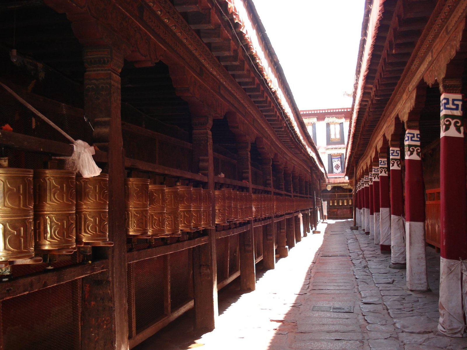 A long corridor of brass prayer wheels under carved wooden eaves at the Jokhang Temple in Lhasa, with red pillars on the right