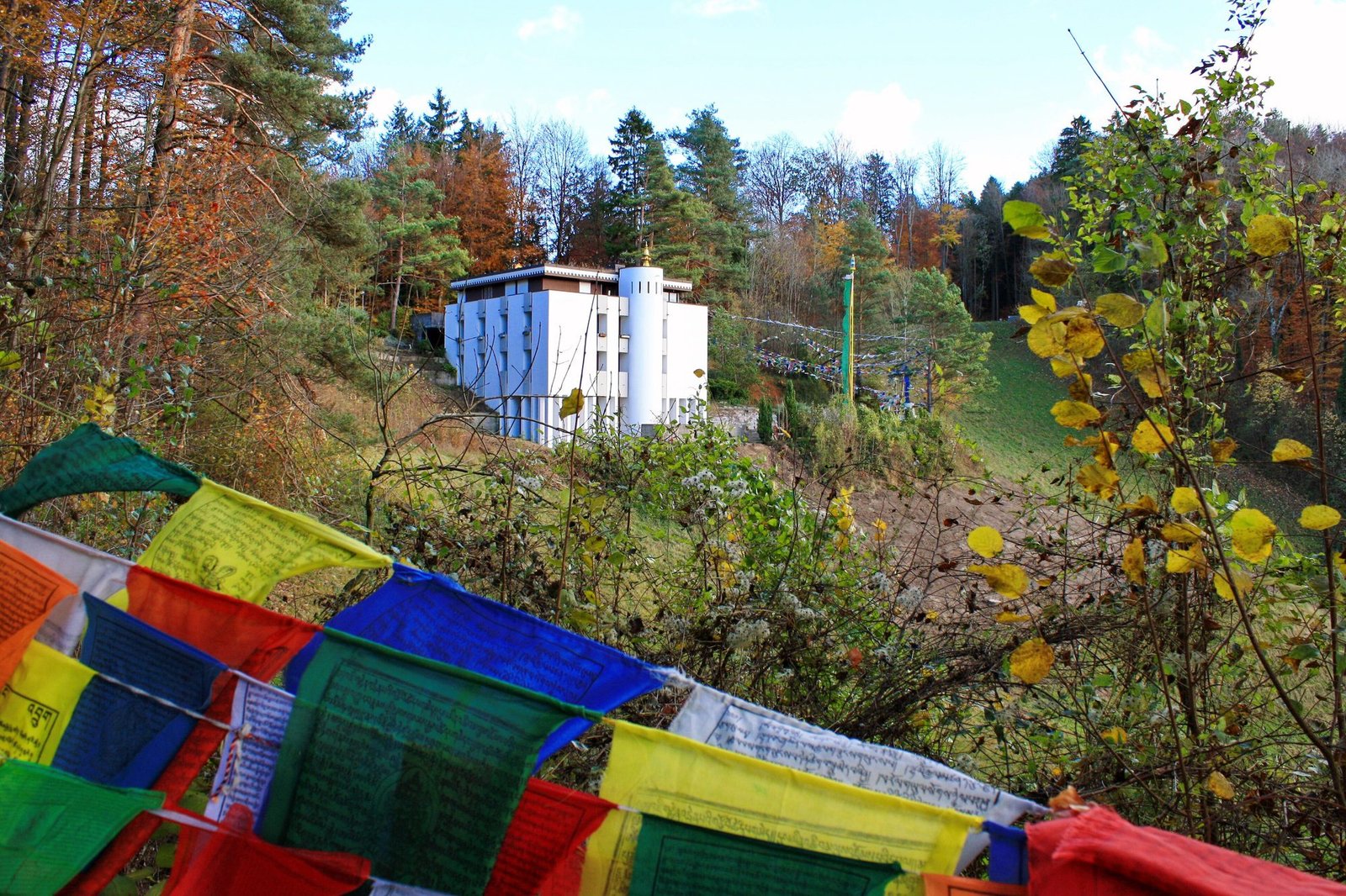 A white monastery building on a wooded hillside in autumn, with prayer flags in the foreground