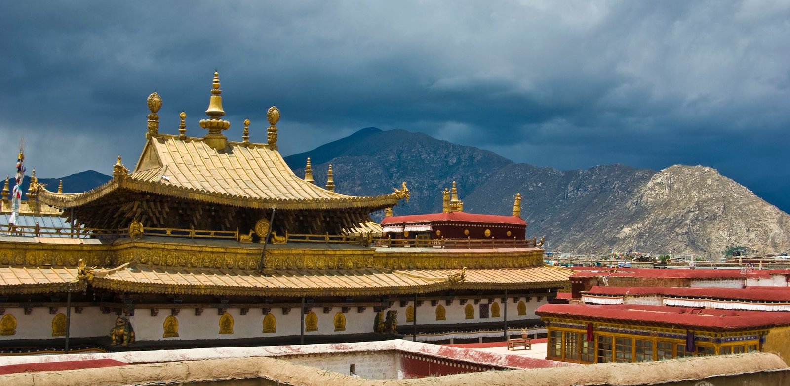 Gilded Tibetan temple roofs with ornaments under a dark stormy sky, mountains behind