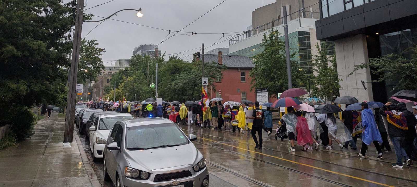 A long column of people with umbrellas, rain ponchos and Tibetan flags marching along a wet city street