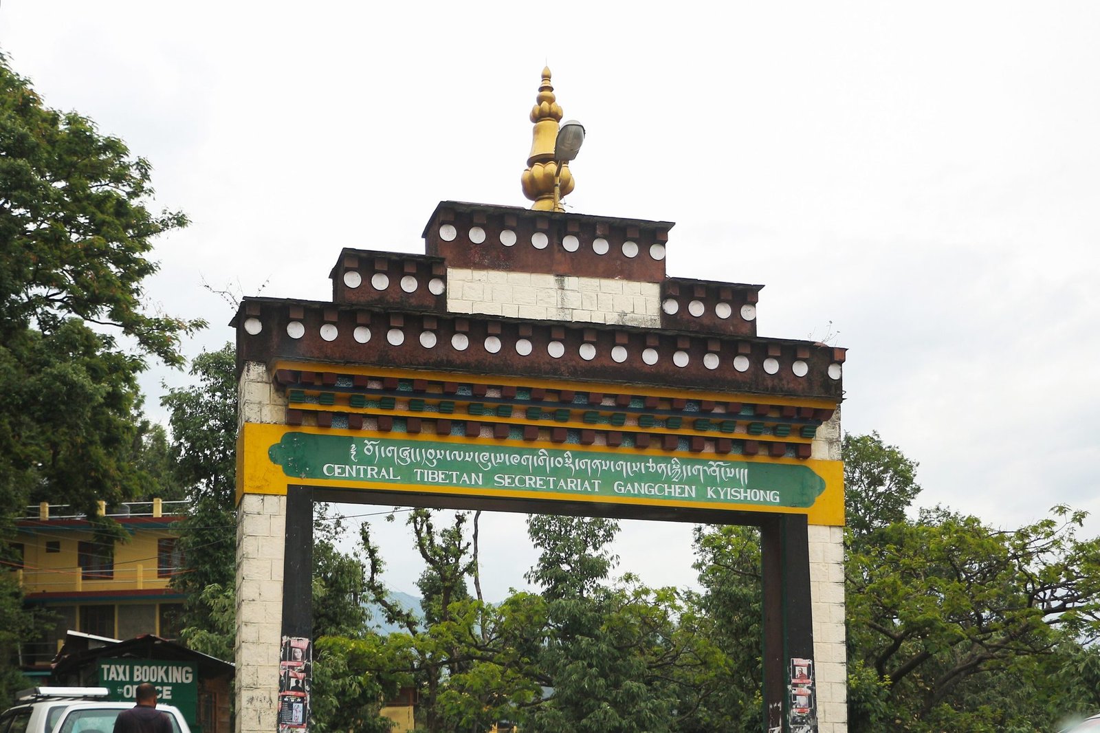 A Tibetan-style gateway with a green sign reading Central Tibetan Secretariat Gangchen Kyishong, trees around it