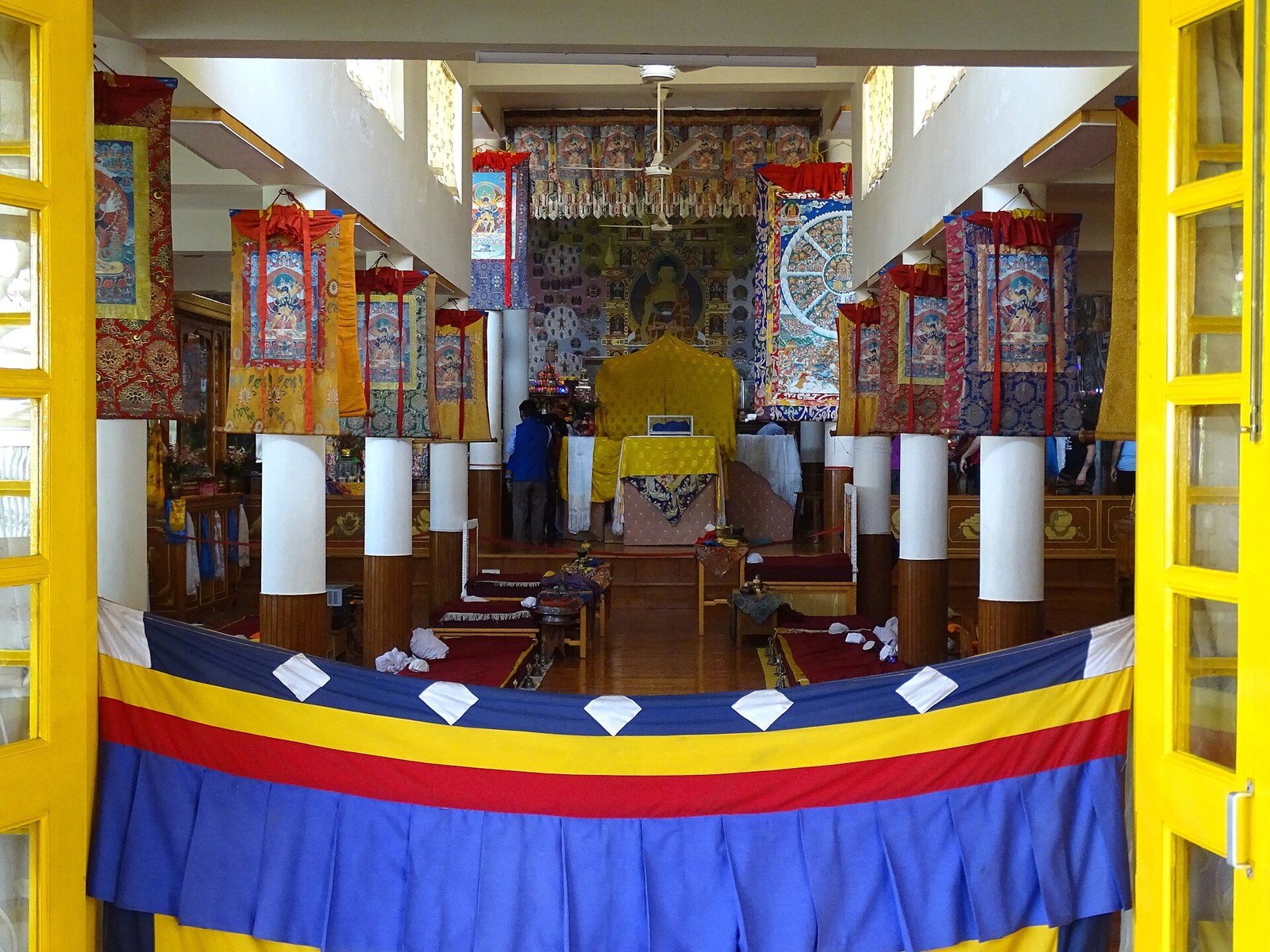 View through open yellow doors into a Tibetan temple hall with painted pillars, hanging thangkas and a throne at the far end