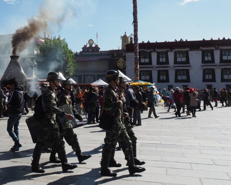 A squad of armed police in helmets and camouflage marching across a sunlit square, pilgrims and a temple behind