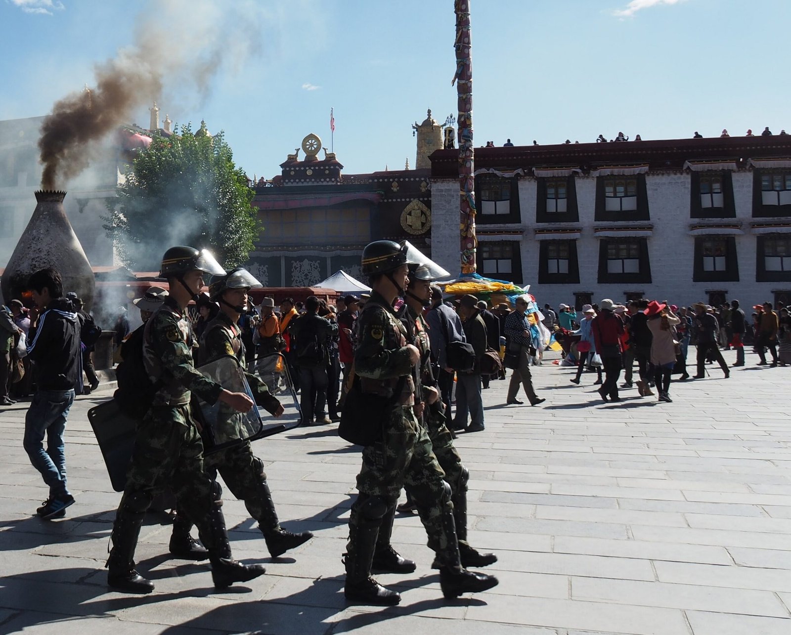 A squad of armed police in helmets and camouflage marching across a sunlit square, pilgrims and a temple behind