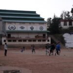 A school hall with the painted words Come to learn, go to serve, above a dirt sports ground where children play in the distance