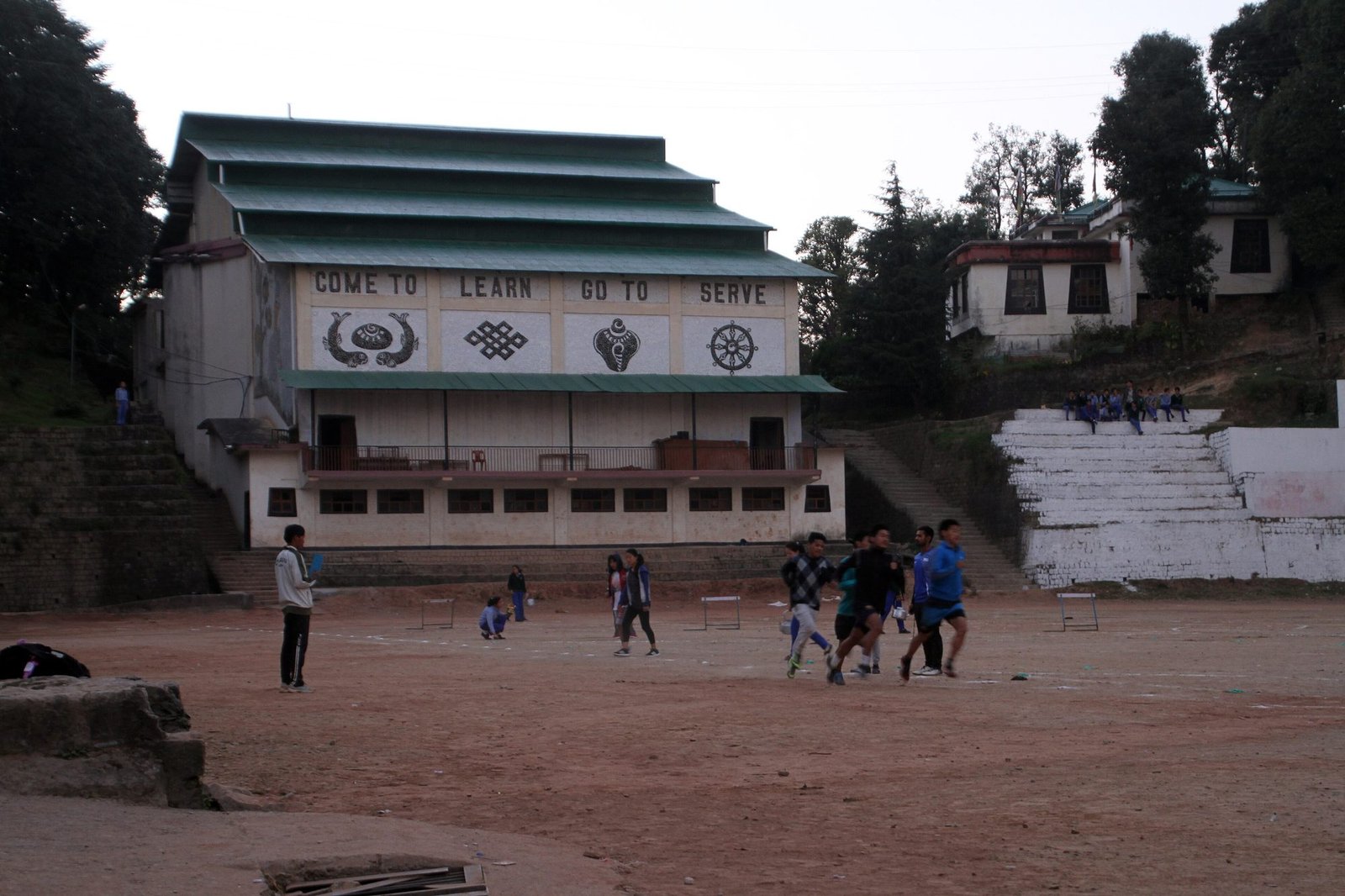 A school hall with the painted words Come to learn, go to serve, above a dirt sports ground where children play in the distance