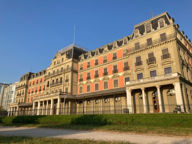 A large 19th-century building with a pink and cream stone facade behind a lawn and railings