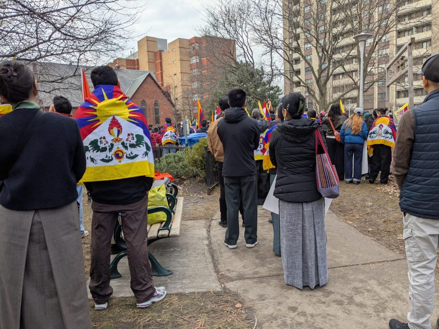 People seen from behind, some wrapped in Tibetan flags, stand in a city park listening to a rally