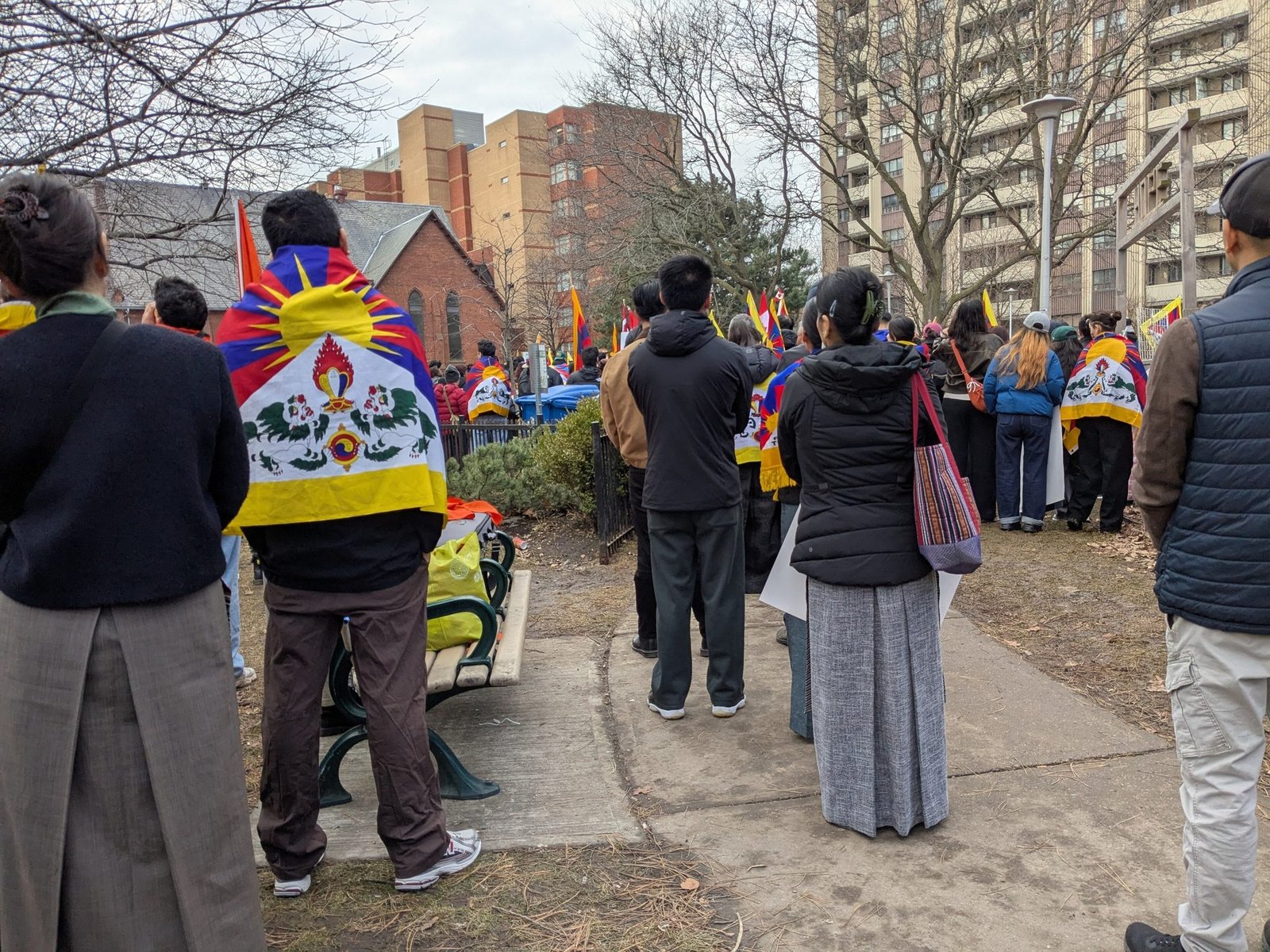 People seen from behind, some wrapped in Tibetan flags, stand in a city park listening to a rally